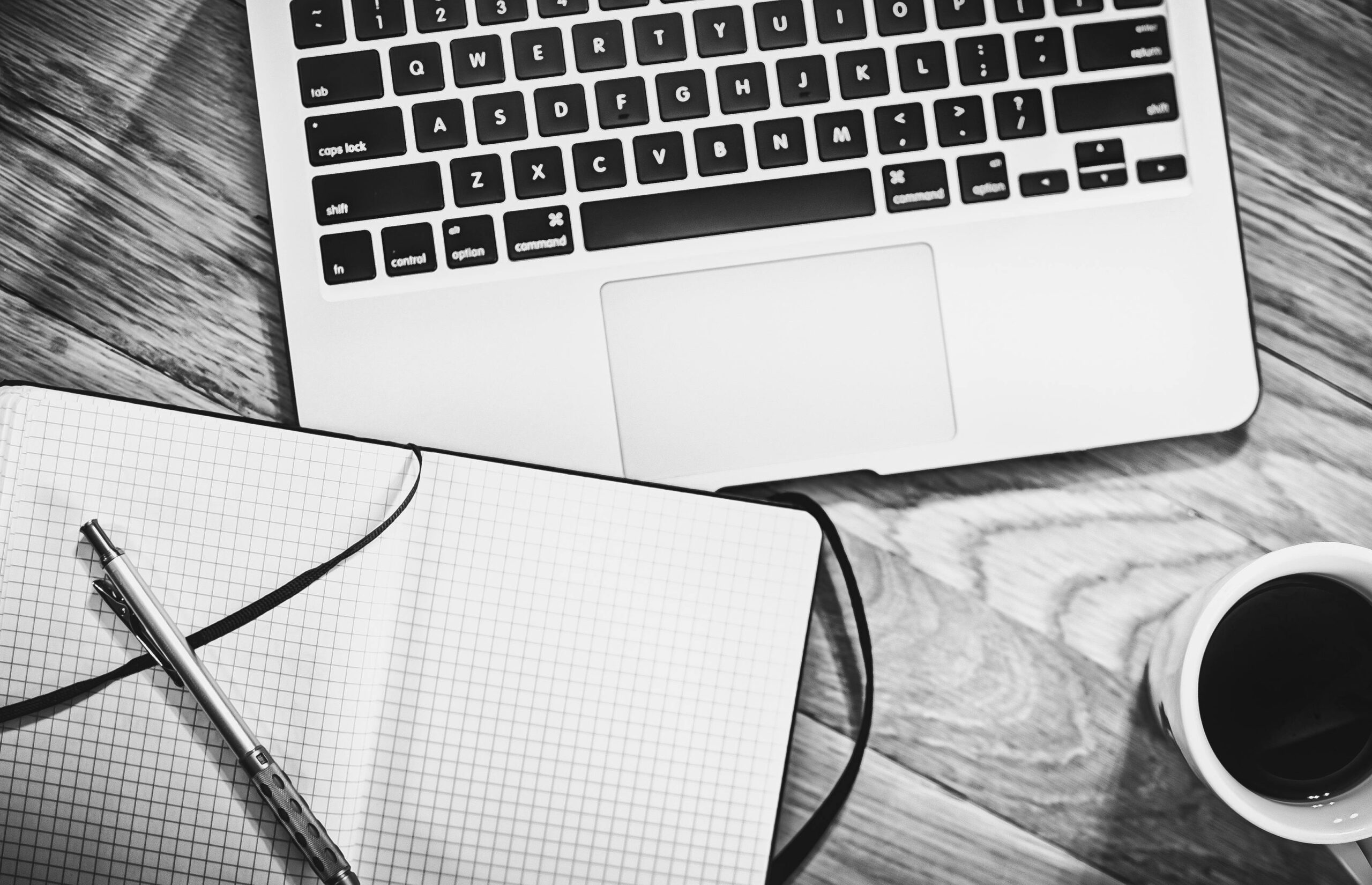 Black and white image of a workspace featuring a laptop, notebook, pen, and coffee cup.